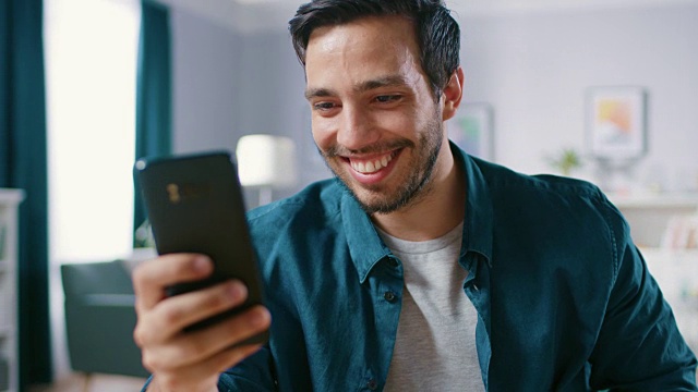 英俊微笑的男人在客厅放松时使用智能手机。男人在网上浏览，使用社交网络，在家里玩。视频素材