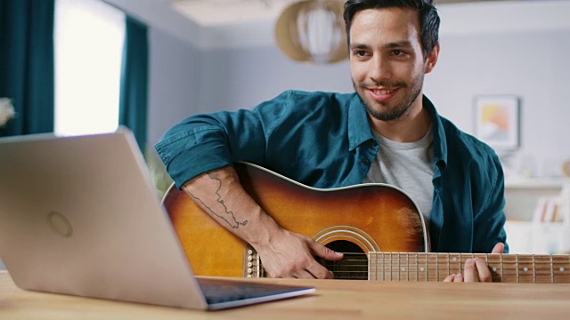 一位才华横溢的年轻人坐在书桌前弹吉他，用笔记本电脑读和弦，写在线视频博客。男人在家练习吉他音乐。视频下载