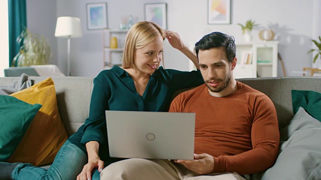 快乐的女朋友和男朋友坐在家里的沙发上，用笔记本电脑，浏览互联网，做电子购物，使用流媒体服务。幸福家庭在舒适的客厅。视频素材
