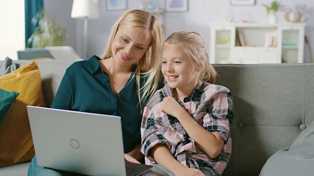 年轻漂亮的妈妈和她可爱的小女儿坐在沙发上使用笔记本电脑。家庭花时间一起看视频和卡通在电脑上。视频素材