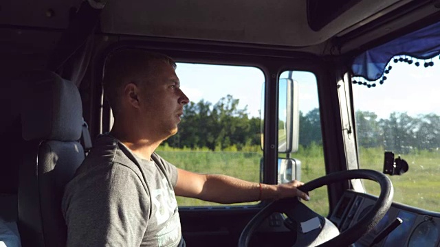 侧面的男子手握方向盘和驾驶卡车通过农村。卡车司机坐在汽车的方向盘旁。一个白人仔细看了看路。侧视图视频下载