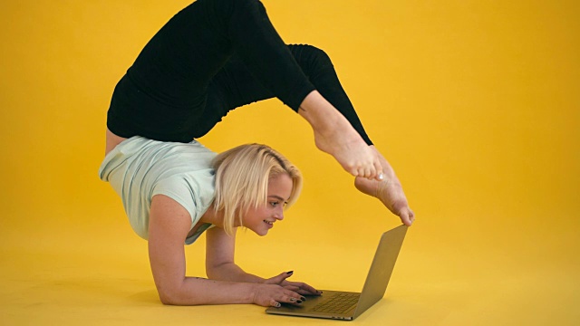 金发模特身材，练习瑜伽倒立，黄色背景下使用笔记本电脑视频下载