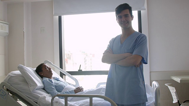 护士交叉双臂微笑地看着镜头，病人躺在医院的床上作为背景视频素材