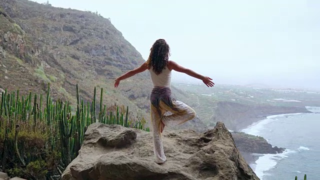 年轻女子在山上做瑜伽，单腿站着俯瞰大海。视频下载