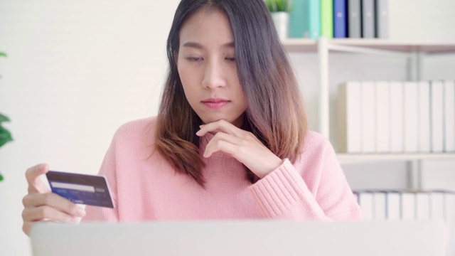 美丽的亚洲女人使用电脑或笔记本电脑购买网上购物的信用卡，而穿着毛衣坐在家里客厅的桌子上。生活方式女性在家的概念。视频素材