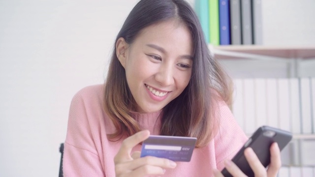 美丽的亚洲女人使用智能手机在网上购物通过信用卡，而穿着毛衣坐在家里客厅的桌子上。生活方式女性在家的概念。视频素材