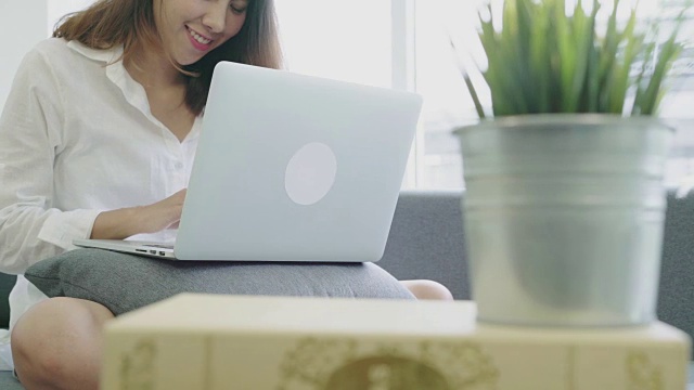 年轻有魅力的亚洲女性或学生坐在客厅的沙发上使用笔记本电脑，感觉放松。视频素材