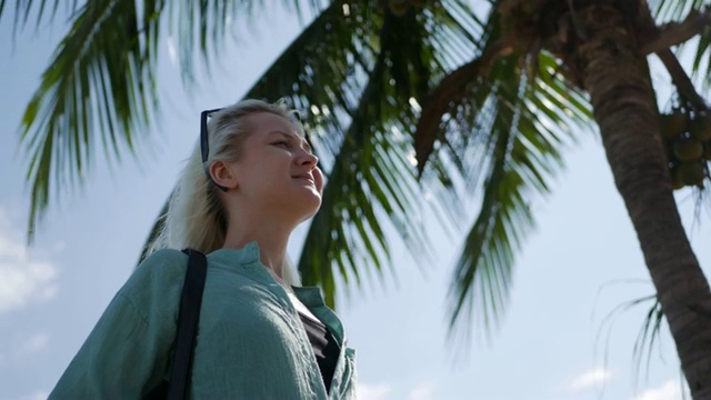 年轻幸福的白人妇女的特写与长金发在太阳镜和绿色衬衫站在棕榈树附近在蓝天的背景。旅游的概念视频素材