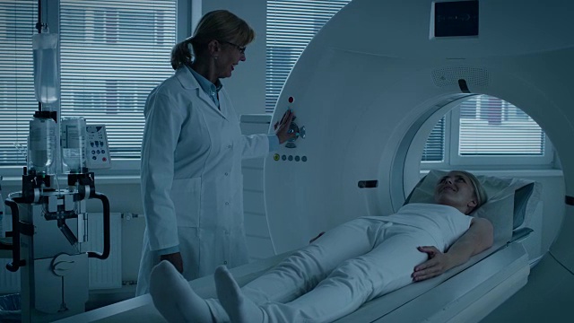在医学实验室女性放射学家控制MRI或CT或PET扫描与女性病人正在进行的程序。医生使用先进的医疗技术进行紧急扫描。蓝色的基调。视频素材