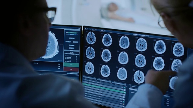 在控制室，医生和放射科医生讨论诊断，同时观看程序和显示器显示的大脑扫描结果，在背景患者接受MRI或CT扫描程序。视频素材