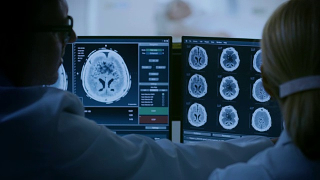 在控制室，医生和放射科医生讨论诊断，同时观看程序和显示器显示的大脑扫描结果，在背景患者接受MRI或CT扫描程序。视频素材