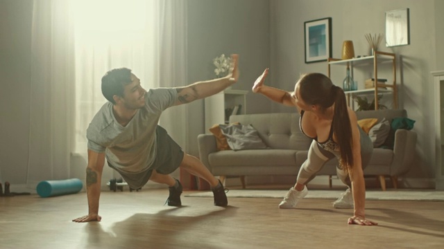 一个强壮美丽的运动健身夫妇穿着运动服在明亮宽敞的客厅里做俯卧撑和互相击掌的慢动作镜头。视频素材