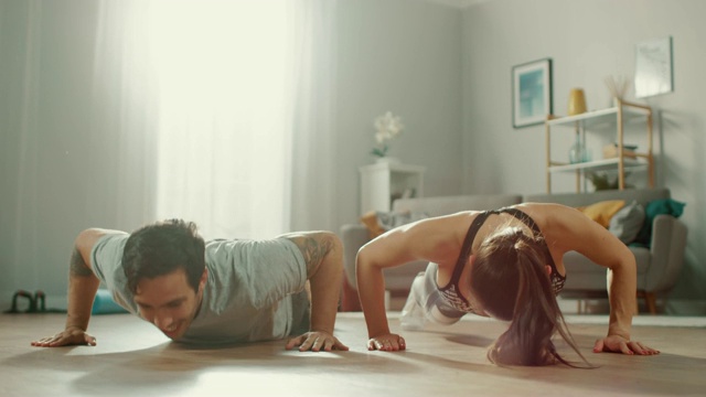 强壮美丽的运动健身夫妇穿着运动服在明亮宽敞的客厅里做俯卧撑，互相击掌。视频素材