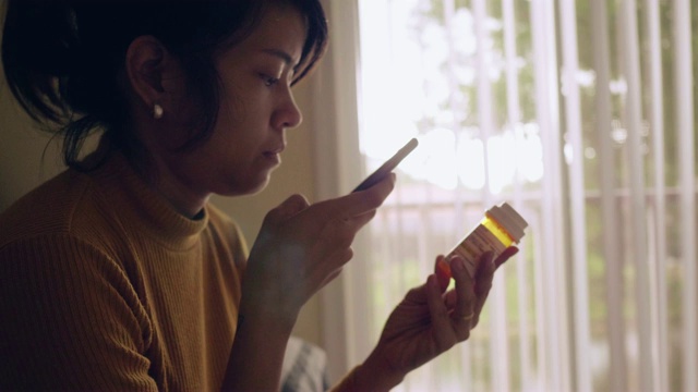 年轻的亚洲女人拿着药瓶，在卧室里讲手机视频素材