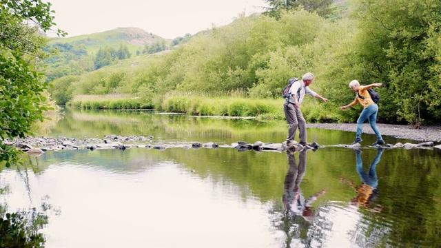 在英国湖区徒步旅行时用垫脚石过河的老年夫妇的慢动作镜头视频下载