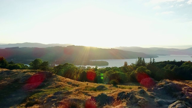无人机拍摄起伏的绿色山丘，树木和温德米尔湖在阳光与镜头光晕，湖区，英国视频下载
