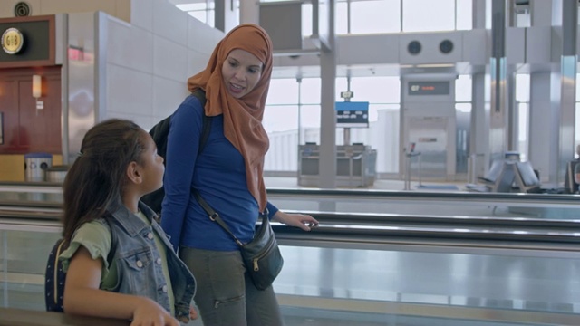 穆斯林母亲戴着头巾，小女儿走在机场候机楼的人行道上。跟踪拍摄。视频下载