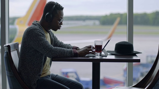 一名混血千禧一代男子在机场航站楼的窗口边听音乐边用平板电脑工作。视频下载