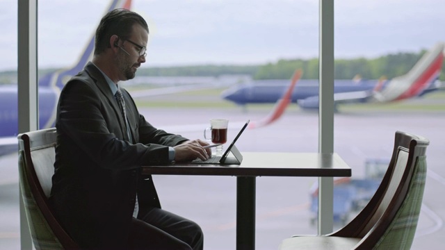 一名商人在机场候机厅咖啡厅的窗口边忙着平板电脑。视频下载