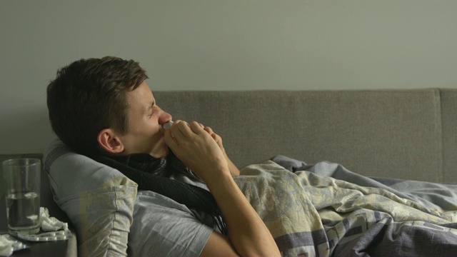 一名男子躺在病床上，旁边是他的药品，用手帕擤鼻子视频素材