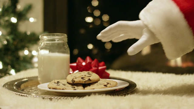 圣诞前夜，圣诞老人戴着手套的手试图从一个托盘里拿起一块巧克力饼干，托盘上放着一杯牛奶，但一只手在圣诞树和壁炉的背景下把它拍走了视频下载