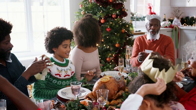 混合种族，多代家庭坐在他们的圣诞餐桌上阅读笑话，戴上纸派对帽从圣诞爆竹，近视频素材