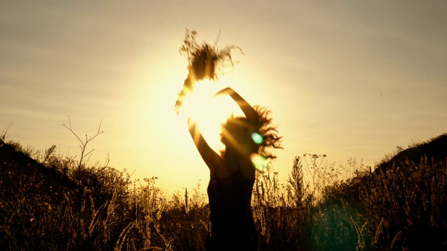 明亮的日落背景上一个女孩的黑色剪影。她在山与山之间的田野里跳着，挥着她的手。底部视图。视频下载