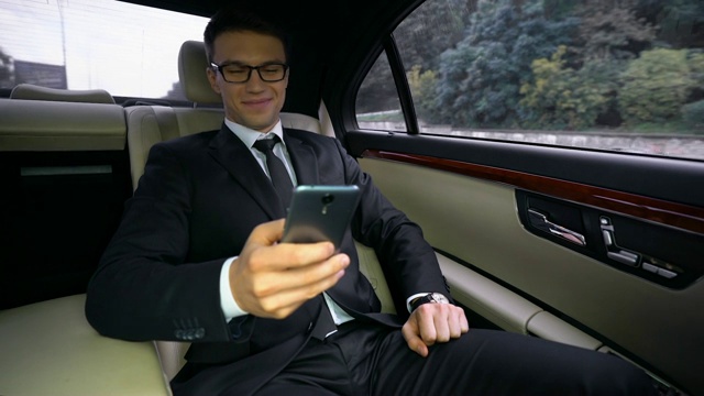 微笑帅气的商人坐在豪华轿车里，在手机上读着好消息视频下载