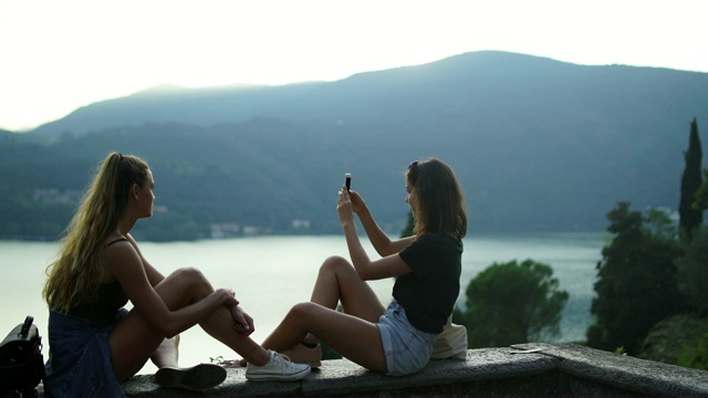 两个女孩在石墙上用智能手机拍照，看下面的湖和山视频素材