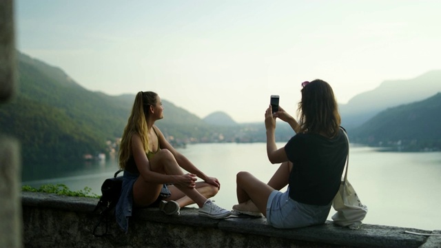 两个女孩在石墙上用智能手机拍照，看下面的湖和山视频素材