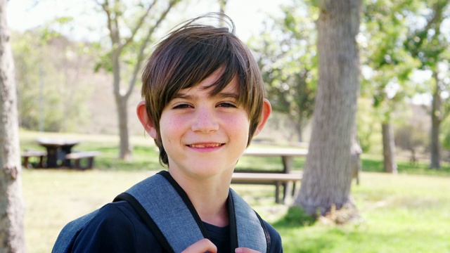 在公园里背着背包对着镜头微笑的小男孩的慢动作肖像视频下载