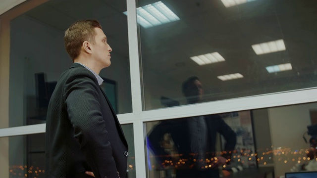 后视图的男企业家穿着公司西装看着窗外，而等待着商业伙伴在现代时尚的晚上办公室与玻璃窗户。自信自豪的CEO在建设视频素材