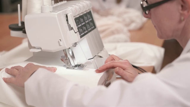 一位女裁缝在缝纫机上缝制白色织物的近距离的侧面视图。另一个裁缝正坐在桌子旁，把珠子缝在纱布上，准备做一件婚纱视频素材