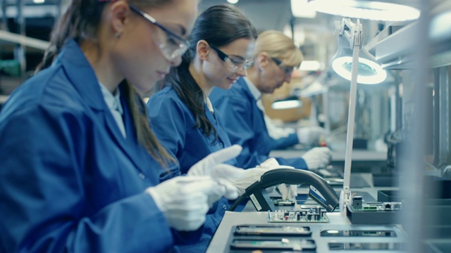 穿着蓝色工作服和防护眼镜的电子厂女工正在用镊子组装智能手机的印刷电路板。拥有更多员工的高科技工厂。视频素材