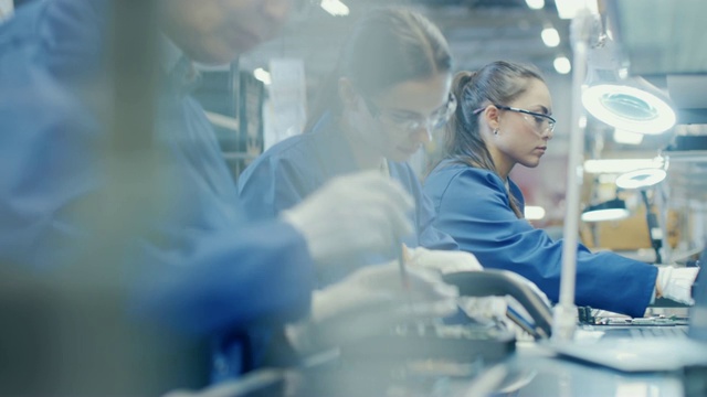 电子工厂里穿着蓝色工作服戴着防护眼镜用镊子组装智能手机的女工。拥有多名员工的高科技工厂设施。视频素材