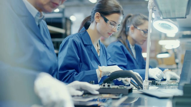 电子工厂里穿着蓝色工作服戴着防护眼镜用镊子和螺丝刀组装智能手机的女工。拥有多名员工的高科技工厂设施。视频素材