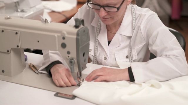 一名女裁缝坐在工作室的桌子旁，开始在缝纫机上缝白色的布料。视频素材