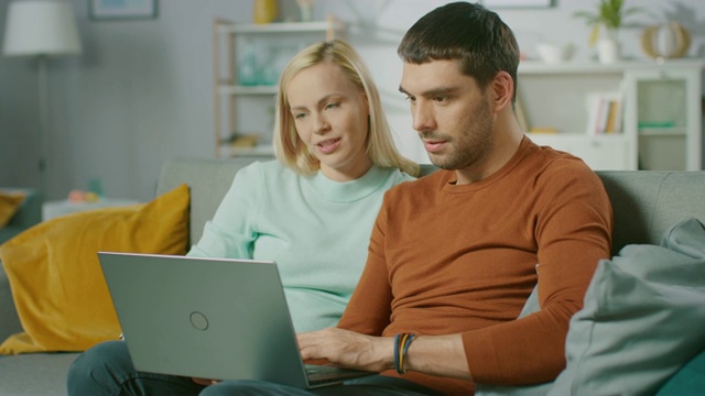 一对可爱的年轻夫妇坐在家里的沙发上，他们使用笔记本电脑，玩得很开心。这是一对美丽幸福的年轻情侣的肖像。视频素材