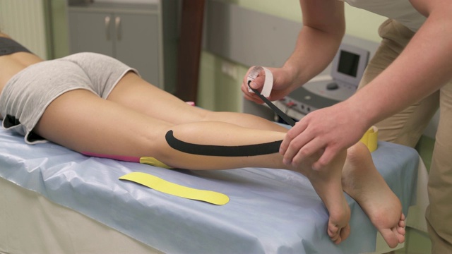 一位男性理疗师拿着一根黑色的运动胶带，把它固定在脚后跟上，应用到运动胶带的跟腱上，然后用纸背摩擦它。视频下载