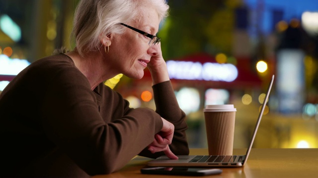 一个快乐成熟的女人晚上坐在城市街道的露天咖啡馆里用笔记本电脑视频素材