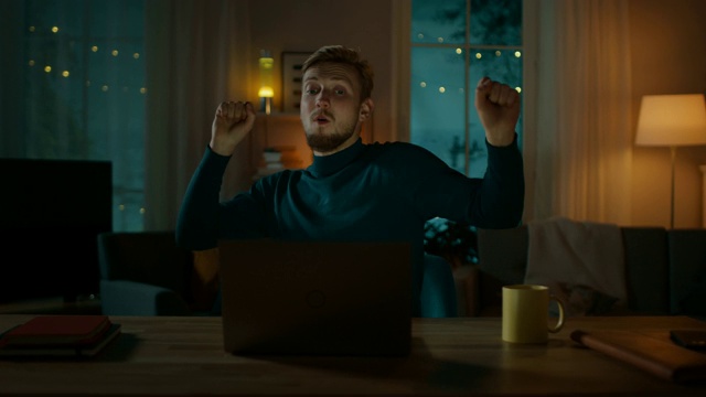 晚上:快乐的年轻人坐在书桌前跳舞，他使用笔记本电脑，听着音乐。视频素材