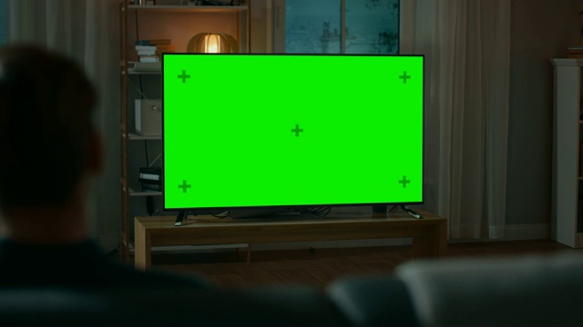 晚上，一名男子坐在家里的沙发上看绿色模拟屏幕电视。温馨的客厅和温暖的灯光。过肩射。视频素材