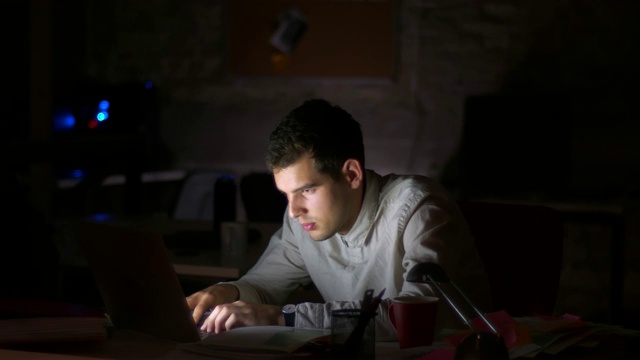 一名友善的白人男子厌倦了在笔记本电脑上打字，坐在漆黑的办公室里，在屏幕旁边睡着了视频下载