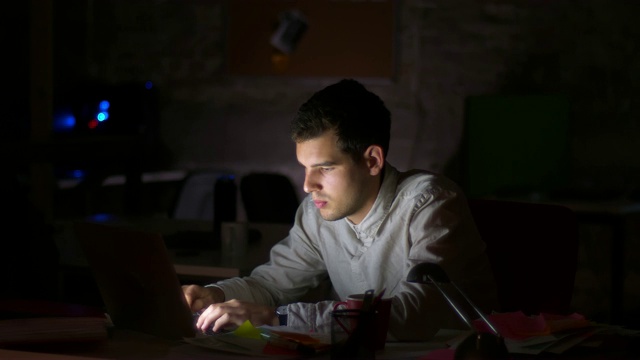集中的白人男性看着电脑屏幕，在键盘上打字，在桌面上工作，深夜，黑暗的背景视频下载
