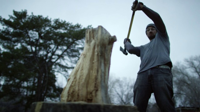 一个戴着编织帽和安全眼镜的四十多岁的白人男子，在一个阴天的黄昏，用斧头把一根被树木环绕的木头劈成两半当柴用视频素材