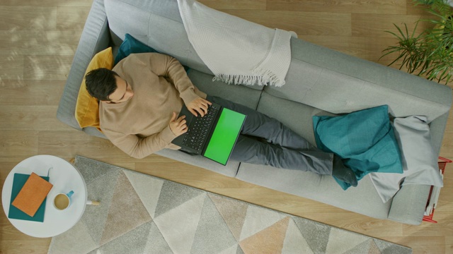 穿着棕色毛衣和灰色牛仔裤的年轻人正躺在沙发上，使用绿色屏幕的笔记本电脑。舒适的客厅与现代室内植物，桌子和木地板。自顶向下。视频素材