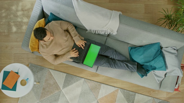 穿着棕色毛衣和灰色牛仔裤的年轻人正躺在沙发上，使用绿色屏幕的笔记本电脑。舒适的客厅与现代室内植物，咖啡桌和木地板。顶部视图与缩小。视频素材