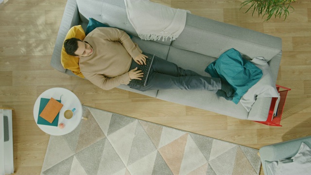 一个穿着棕色毛衣和灰色牛仔裤的年轻人正躺在沙发上，用着笔记本电脑。舒适的客厅与现代室内植物，咖啡桌和木地板。Top View with Close Up。视频素材