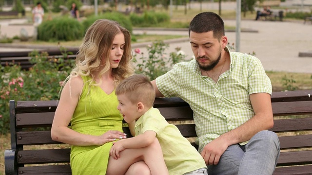 快乐的年轻家庭和他的儿子在公园的长椅上休息在夏天。视频下载