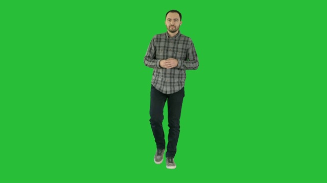 年轻自信的男人，穿着衬衫和牛仔裤走向相机，并指着一边的绿色屏幕，色度键视频素材